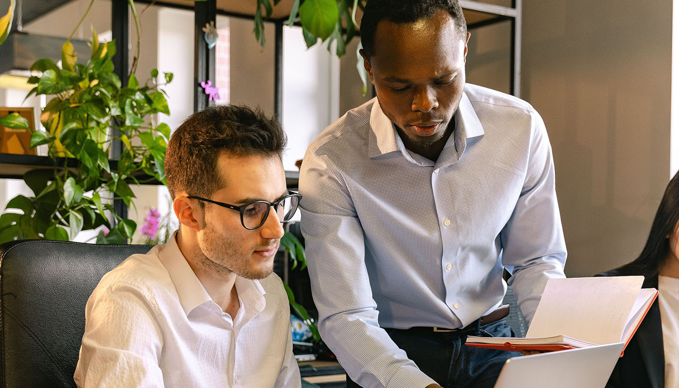 Three colleagues are gathered around a laptop in a modern office, engaged in a focused discussion. One person stands and gestures toward the screen while the others listen and take notes, suggesting collaboration on a project.