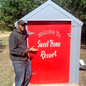 Man standing in front of a red shed, smiling, and gesturing.