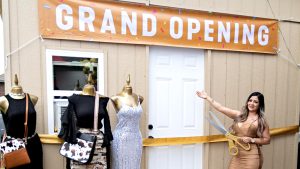 This is an image of a person posing in front of a storefront with a grand opening sign, and there is a store sign above a display of mannequins wearing dresses.