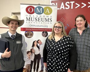 The image is a collage of two photographs. In the top photo, two people are wearing cowboy hats and posing with the sign "Oklahoma Museums Association." The bottom photo features a person standing in front of the same sign, which includes a banner. The person in the bottom photo is holding a book.