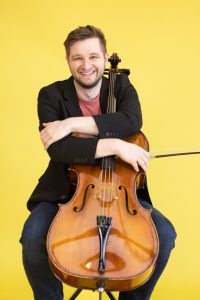 A man is sitting with a cello, smiling, and leaning on it.