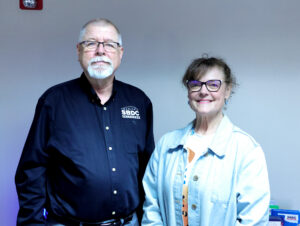 The image shows two people, likely a man and a woman, standing side by side. The man is wearing a black shirt and glasses, and the woman is wearing a blue shirt. They both appear to be older adults. In the background, there is a table with some items on it and what looks like a wall or a decorative element.