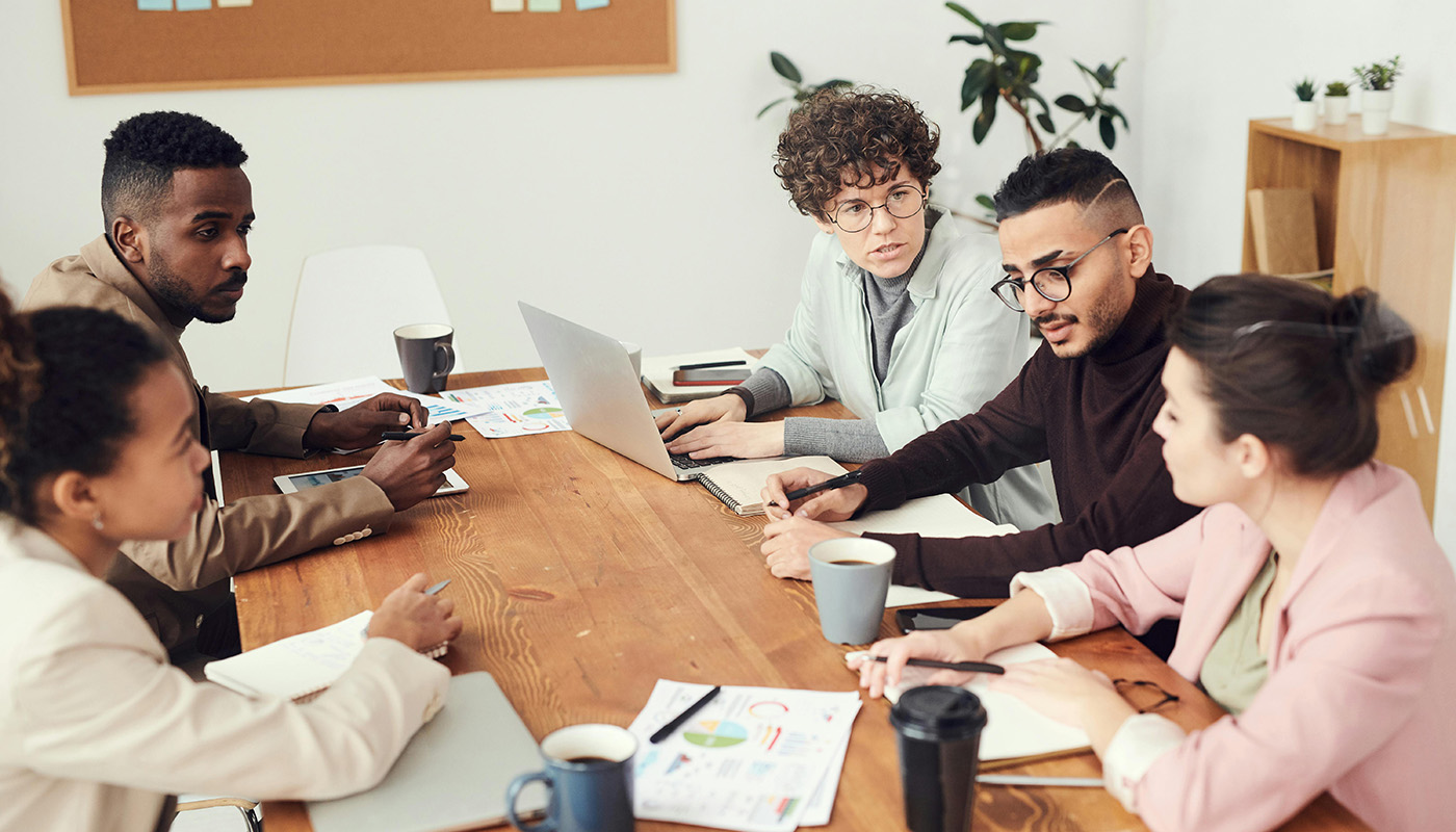 A group of professionals collaborate around a table, discussing documents and working on laptops in an office setting.