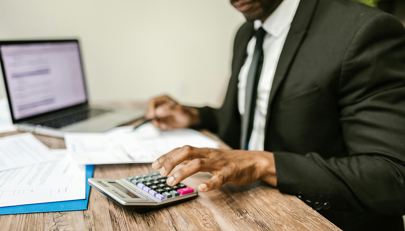 A business professional in a suit uses a calculator at a desk with paperwork and a laptop, reviewing financial documents related to small business taxes.
