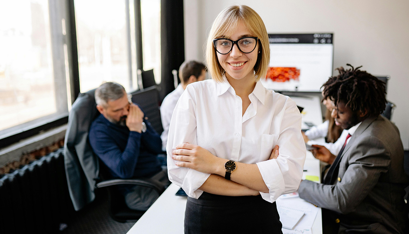 A woman wearing glasses and a white blouse stands with her arms crossed in a modern office, smiling at the camera while coworkers collaborate at computers in the background.