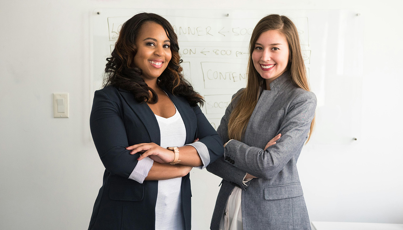 Two professional women in business attire stand side by side with arms crossed, smiling confidently in front of a whiteboard.