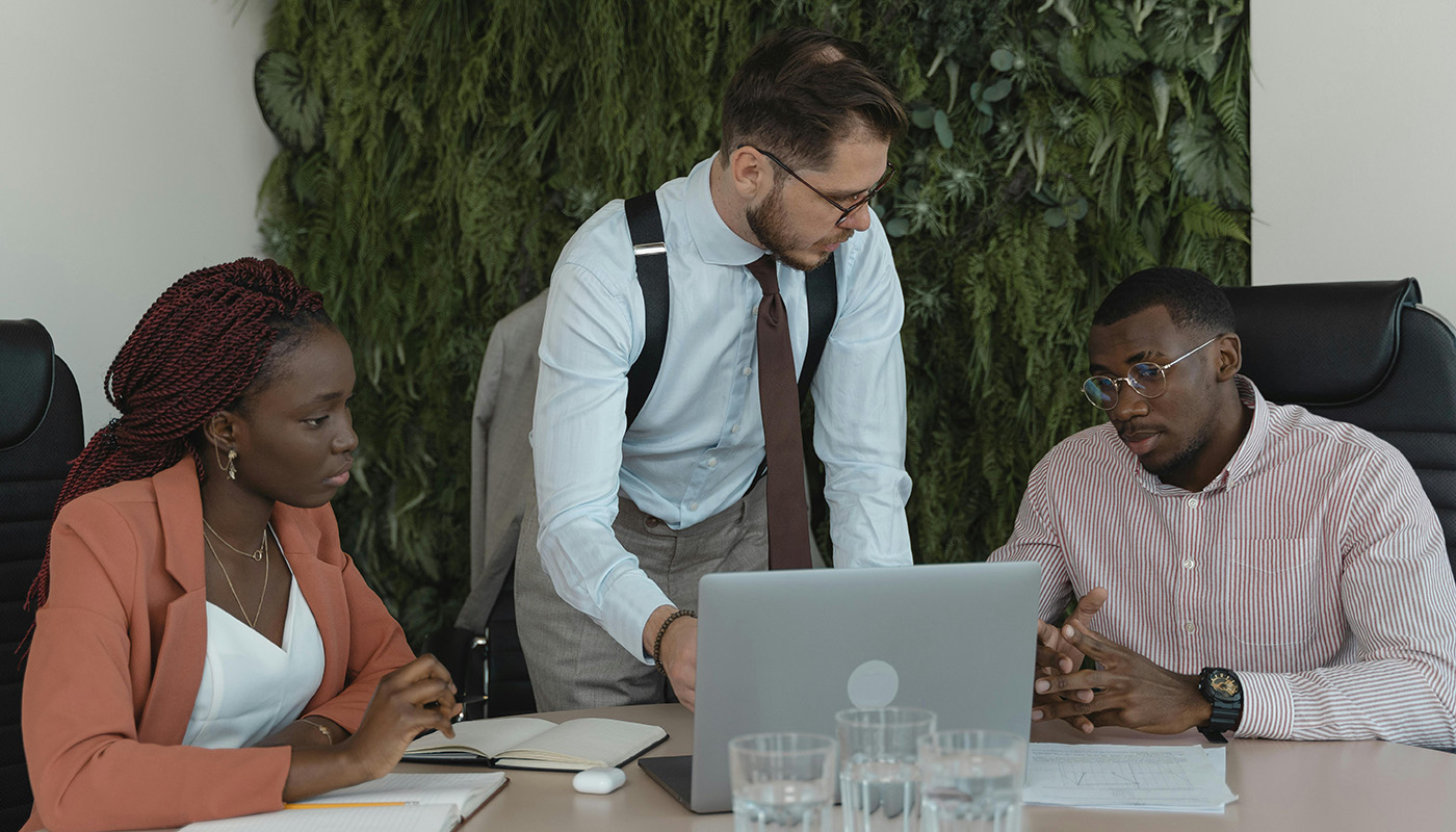 Three colleagues are gathered around a laptop in a modern office, engaged in a focused discussion. One person stands and gestures toward the screen while the others listen and take notes, suggesting collaboration on a project.
