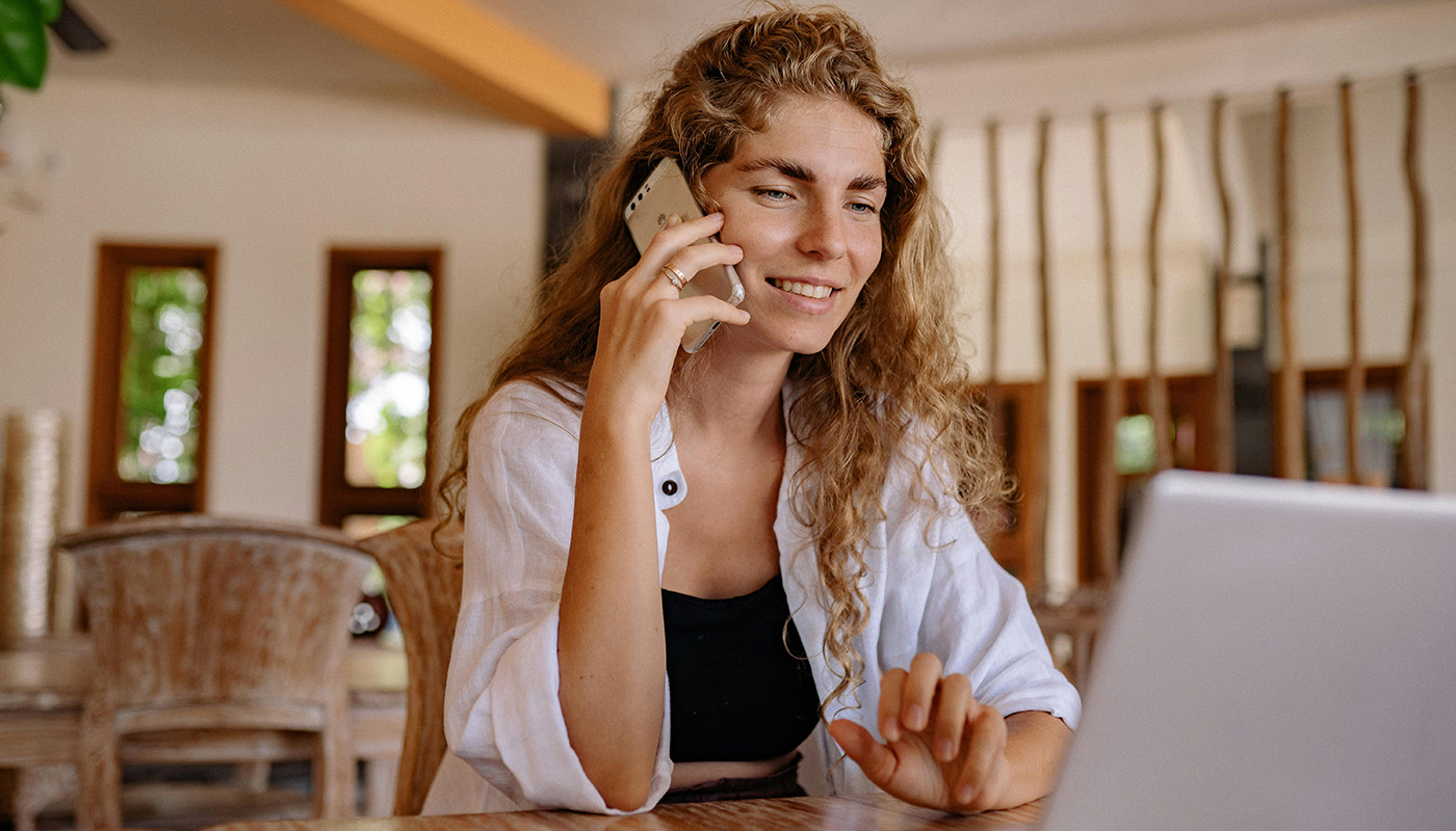 A woman with curly hair smiles while talking on her phone and using a laptop at a wooden table in a bright, cozy room.