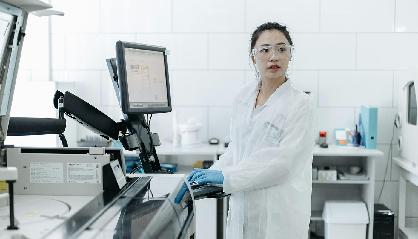 Three colleagues are gathered around a laptop in a modern office, engaged in a focused discussion. One person stands and gestures toward the screen while the others listen and take notes, suggesting collaboration on a project.