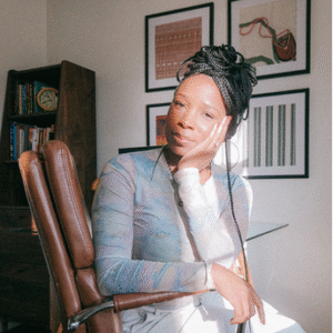 In the center of a room with white walls, there is a woman sitting on a brown leather chair. She has her hair styled in braids and is wearing a blue shirt that matches the color of the chair she's sitting on. The chair appears to be an office chair, suggesting it might be used for work or study purposes. Behind the woman, there are several framed pictures hanging on the