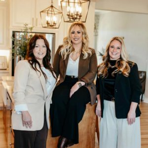 In the image, there are three women standing in a kitchen. The woman on the left is wearing a brown blazer and has long hair that falls over her shoulders. In the center of the group, another woman is wearing a black blazer with gold accents. To the right, the third woman is also dressed in a black blazer but without any accessories or additional clothing items.