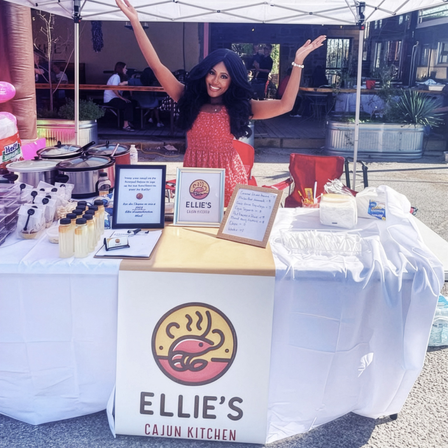 Oklahoma SBDC client Ellie Daniels, owner of Ellie’s Cajun Kitchen, stands smiling with her arms raised behind a vendor table at an outdoor event. The table displays Ellie’s Cajun Kitchen signage, food containers, bottled products, and serving supplies beneath a canopy.