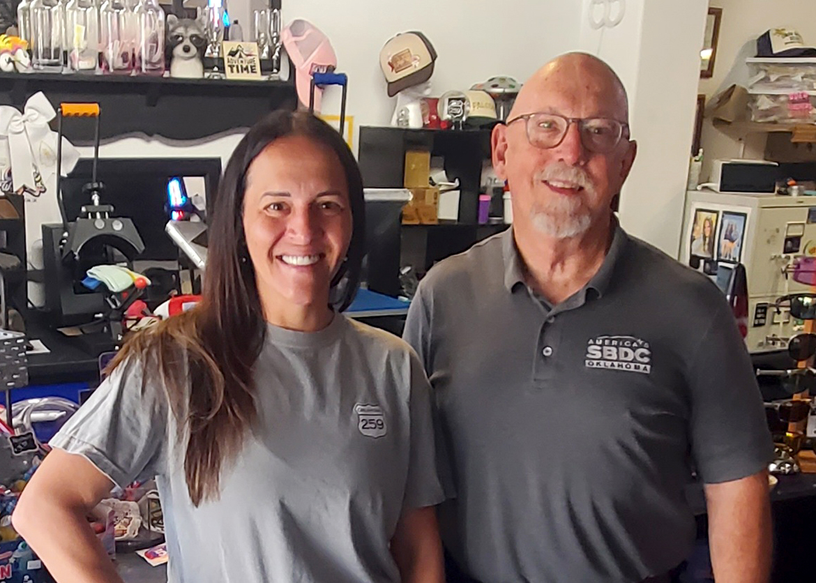 Jessica Shank, owner of Camp 259 T-Shirt & Souvenirs, stands with Oklahoma SBDC Business Advisor Joe Silk inside the business. Both are smiling at the camera, with apparel, hats, printing equipment, and other merchandise visible in the background.