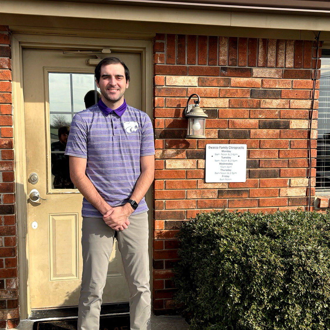 Oklahoma SBDC client Coleton Oakes, owner of Owasso Family Chiropractic, stands smiling outside the entrance to his chiropractic office. Oakes is wearing a purple striped polo and stands beside a brick wall displaying the Owasso Family Chiropractic office hours.